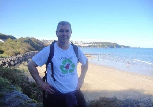 Steve is wearing a rucksack, standing with his hands on his waist, with a beach and see in the background