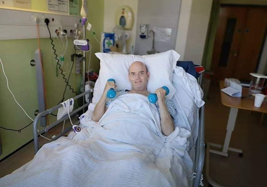 Kev Hill Man laying in a hospital bed, propped up on pillows, holding two blue dumbbell hand weights