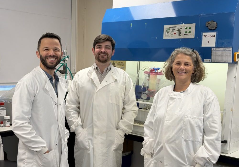 Bettina Wilm and team (1) Three researchers, wearing white lab coats, standing in front of the machine in the laboratory.