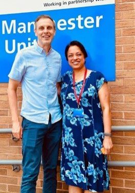 Dr Glen Cooper and Dr Anu Jayanti Glen and Anu standing in front of a brick building with a Manchester University sign on the wall.