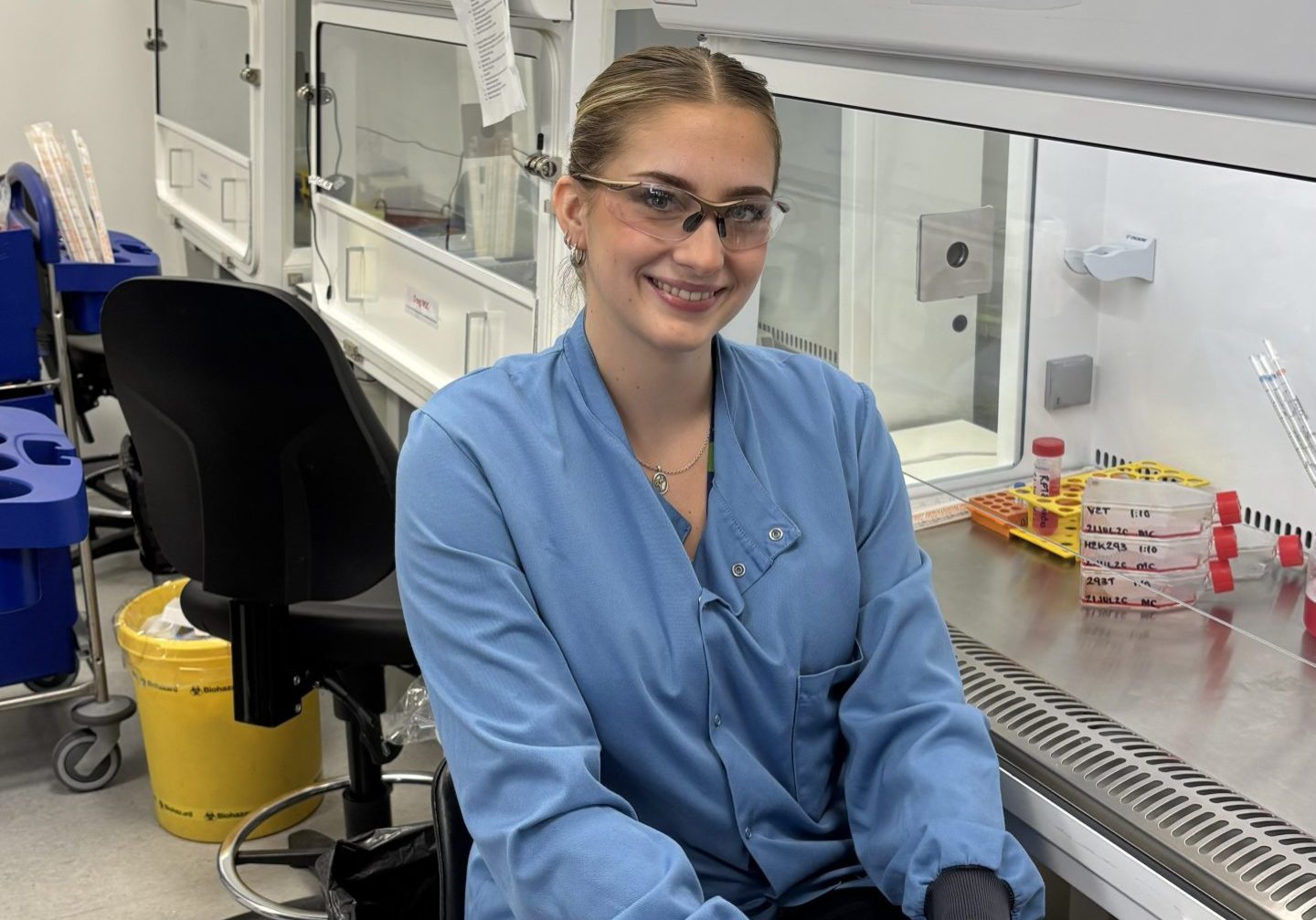 Female PhD student, wearing a blue lab coat, sitting at a research lab bench.